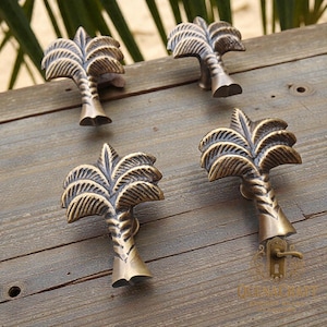 Op de afbeelding: Vier antieke messing palmboomvormige kastknoppen. De knoppen hebben gedetailleerde getextureerde bladeren en stammen, gemonteerd op een verweerd houten oppervlak. Een decoratief hardware-element.