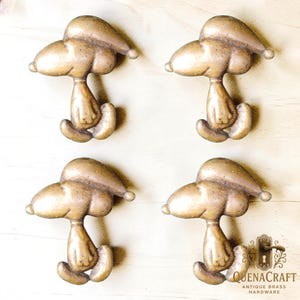 May include: Four antique brass hardware pulls in the shape of a cartoon character. Each pull is a light bronze color with a detailed design. The pulls are arranged on a light wood surface. The text "QUENACRAFT ANTIQUE BRASS HARDWARE" is visible.