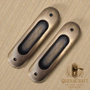May include: A pair of antique brass recessed door pulls. Each pull has an oval shape with a textured, rectangular grip area. The pulls are a warm, aged brass colour and have small screw holes at each end. The QuenaCraft logo is in the bottom right corner.