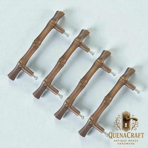 Könnte beinhalten: Vier Schrankgriffe im Bambus-Stil mit warmbraunem Finish. Jeder Griff hat ein Bambusstangen-Design mit einem Quersteg an jedem Ende und einer silbernen Schraube zur Befestigung. Das Logo "QUENACRAFT ANTIQUE BRASS HARDWARE" ist sichtbar.