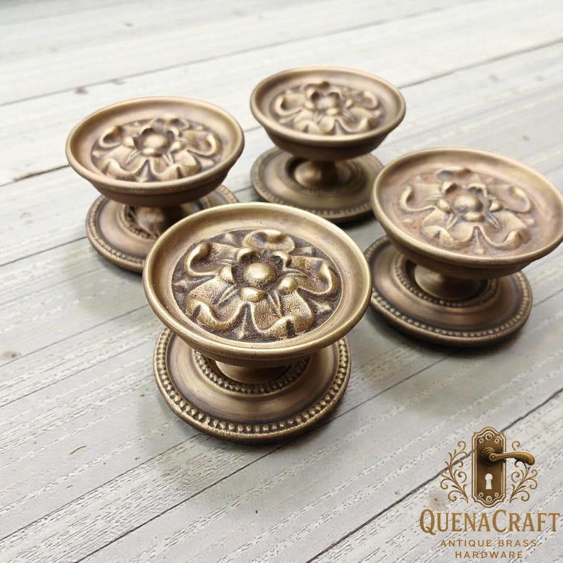May include: Four antique brass door knobs with a floral design. The knobs have a round base with a beaded edge and a detailed flower pattern on the top. The knobs are a warm, aged brass color.