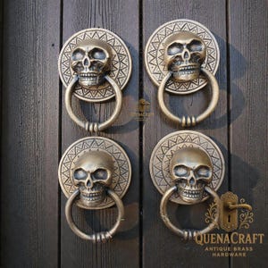May include: Four antique brass door knockers with skull designs. Each knocker features a skull with a circular patterned backplate and a ring handle. The knockers are mounted on a dark wooden door. The brand name "QUENACRAFT ANTIQUE BRASS HARDWARE" is visible.
