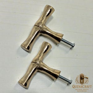May include: Two brass-colored cabinet pulls with a bamboo-like design. Each pull has a T-shape with a screw attached for installation. The pulls are set against a light-colored wooden surface. The logo "QUENACRAFT ANTIQUE BRASS HARDWARE" is in the bottom right corner.