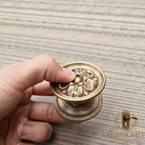 May include: Antique brass door knob with a floral design. The knob is round with a detailed flower pattern on the top and a beaded edge on the base. The hardware is held by a hand.