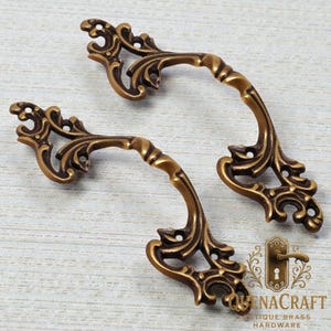 May include: Two antique brass cabinet handles with ornate, swirling floral designs. The handles have a curved shape and a warm, bronze finish. The handles are likely for drawers or cabinets.