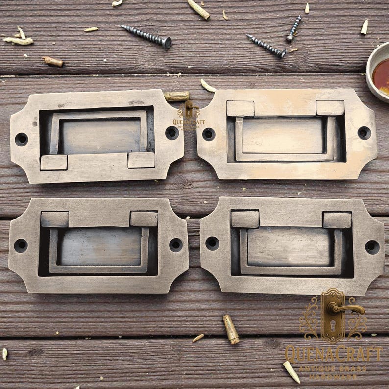 May include: Four antique bronze drawer pulls with a rectangular recessed handle and decorative edges. Each pull has two screw holes for mounting. The pulls are arranged on a weathered wooden surface.