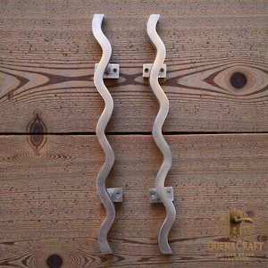 May include: Two silver-colored, wavy door pulls mounted on a wooden surface. The pulls have a unique, elongated S-shape design. Each pull is attached to the wood with small, square mounting plates. The wood has a natural grain pattern.