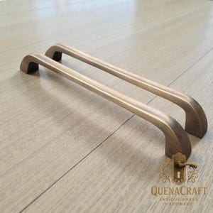 Pode incluir: Duas puxadores de armário de latão antigo com um design curvo. Os puxadores têm uma cor metálica quente e estão colocados sobre uma superfície de grão de madeira clara. O logotipo "QUENACRAFT ANTIQUE BRASS HARDWARE" é visível.