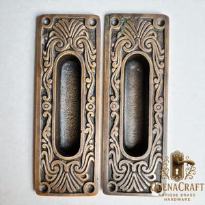 May include: A pair of antique brass hardware pieces featuring an elaborate, swirling design. Each piece is rectangular with an oval recessed handle and decorative floral and swirl patterns. The hardware has an aged, patinated finish.