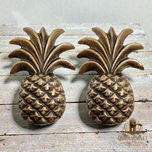 Könnte beinhalten: Zwei bronzefarbene Ananas-Schrankknöpfe. Die Knöpfe haben detaillierte Texturen und ein tropisches Thema. Die Knöpfe sind aus Metall und eignen sich für Möbel oder Dekoration. Die Knöpfe werden auf einer Holzoberfläche präsentiert.