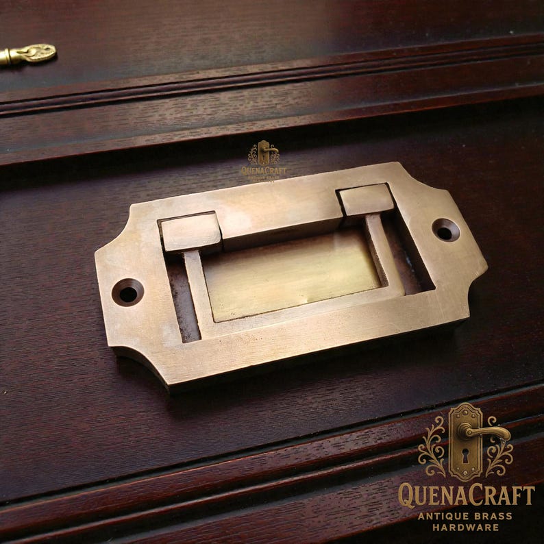 May include: Antique brass drawer pull with a rectangular recessed label area. The hardware has a rectangular shape with rounded edges and two screw holes. The brass has a warm, aged patina. The logo "QUENACRAFT ANTIQUE BRASS HARDWARE" is visible.