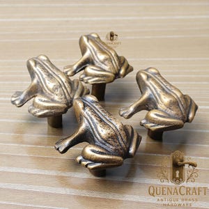 May include: Four antique brass frog-shaped cabinet knobs. Each knob features a detailed frog design with a bronze finish. The knobs are mounted on short stems, suitable for drawers or cabinet doors. The knobs are from QuenaCraft.