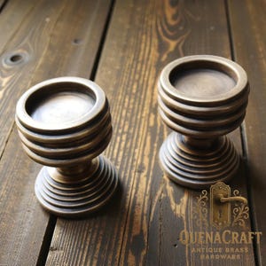 Può includere: Due portacandele antichi in ottone con un design a coste. I supporti hanno una parte superiore e inferiore circolari, con una serie di creste orizzontali che creano un effetto decorativo. La base ha un design a spirale. Il marchio "QuenaCraft Antique Brass Hardware" è visibile.