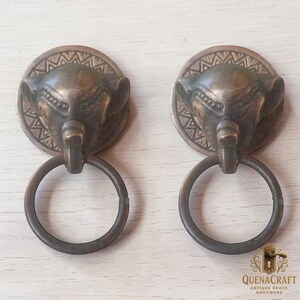May include: Two antique brass elephant head door knockers. Each knocker features a detailed elephant head with a ring pull. The circular backplates have a decorative border. The hardware is a warm bronze color.
