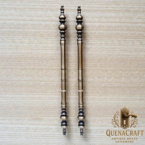May include: Two antique brass door pulls with a bamboo-like design. Each pull features a series of decorative elements, including rounded and cylindrical shapes, and a pointed top and bottom. The pulls are displayed against a light wood grain background.