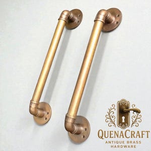 Pode incluir: Duas puxadores de porta de latão antigo. Cada puxador apresenta um tubo reto de latão conectado a um cotovelo curvo e uma placa de montagem circular. O hardware é de uma cor dourada quente. O logotipo QuenaCraft está no canto inferior direito.
