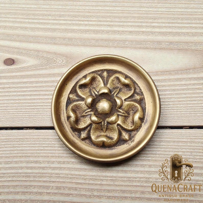 May include: A round, antique brass door knob with a floral design. The knob features a raised, detailed flower pattern in the center, surrounded by a circular border. The finish is a warm, aged gold color, suggesting a vintage aesthetic.