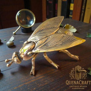 Op de afbeelding: Een gedetailleerde messing vliegensculptuur met ingewikkelde vleugel- en lichaamsdetails. De sculptuur staat op een houten oppervlak, met een vergrootglas op de achtergrond. Het QuenaCraft-logo is rechtsonder zichtbaar.