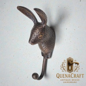 May include: A bronze-toned wall hook in the shape of a rabbit's head. The hook curves downwards with a rounded tip. The rabbit's head features textured fur and a focused expression. The hook is mounted on a light-coloured wall.
