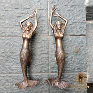 May include: Two bronze mermaid door handles. Each handle features a mermaid with arms raised, a scaled tail, and a decorative necklace. The handles are mounted on a textured, gray surface.