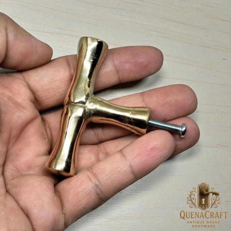 May include: A gold-colored cabinet pull with a T-shaped design. The hardware has a smooth, polished finish and includes a silver screw for installation. The product is from QuenaCraft Antique Brass Hardware.