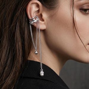 Pode incluir: Punho de orelha prateado com correntes penduradas e pequenos pingentes redondos. O punho de orelha envolve a orelha e apresenta um design decorativo. As correntes pendem, criando um visual elegante e moderno. Uma peça de joalharia moderna.