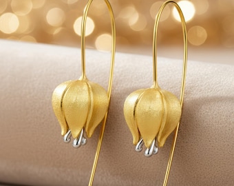 Puede incluir: Un par de pendientes colgantes dorados. Cada pendiente presenta un colgante texturizado en forma de flor con detalles plateados. Los pendientes tienen un diseño de alambre largo y curvado para orejas perforadas. El fondo es suave y dorado.