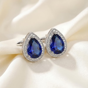 May include: Silver cufflinks featuring a teardrop-shaped, deep blue gemstone centre, encircled by a halo of small, clear stones. The cufflinks are displayed on a soft, white satin background.