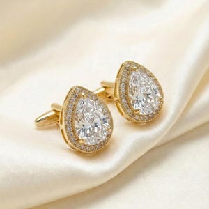 May include: Two gold-coloured cufflinks, each featuring a clear, pear-shaped gemstone at the centre. The gemstones are encircled by a halo of smaller, clear stones. The cufflinks are presented on a cream-coloured, satin-like material.