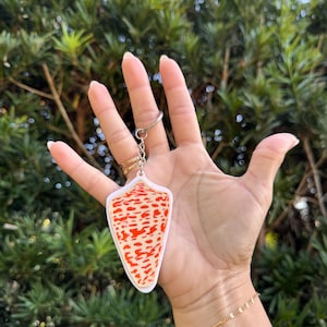 Puede incluir: Un llavero blanco y naranja estampado con un llavero plateado. El llavero tiene un diseño abstracto con un borde blanco. El fondo es una vista borrosa de follaje verde.