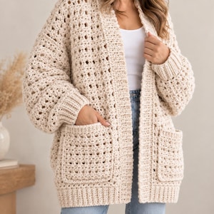 Pode incluir: Um cardigã de crochê cor de creme de corte oversize com padrão de ponto aberto, mangas compridas com punhos canelados, bainha canelada e dois bolsos frontais grandes. A peça é usada sobre uma regata branca e calças de ganga azuis.