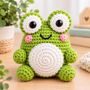 May include: A green crochet frog plush toy featuring large white circular eyes with black pupils, a white circular belly patch, and small pink stitched cheeks. The frog has a simple black embroidered smile and two small arms and legs.