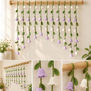 May include: A wall hanging featuring multiple vertical strands of crocheted vines with small green leaves and alternating white and light purple bell-shaped flowers, all suspended from a horizontal wooden rod mounted on a plain wall.