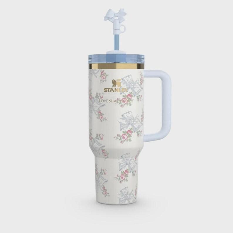 May include: A white insulated tumbler with a handle, featuring a repeating pattern of light blue bows and small pink floral clusters. The Stanley logo and LoveShackFancy text are printed in gold. It includes a clear lid and a white straw with a bow topper.