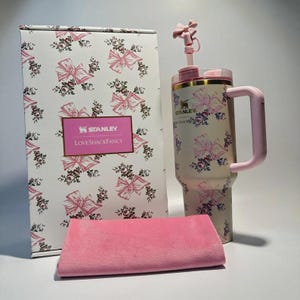 May include: A Stanley LoveShackFancy tumbler with a floral and pink bow pattern, a matching pink handle, and a bow-shaped straw topper. The set includes a rectangular box with the same floral pattern and a pink rectangular fabric cloth.