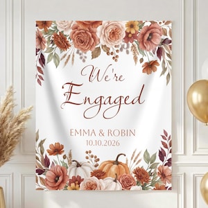 May include: A rectangular white fabric banner featuring the text "We're Engaged" in brown script, followed by "EMMA & ROBIN" and "10.10.2026". The design is framed by watercolor illustrations of orange and white pumpkins, roses, and autumn foliage.