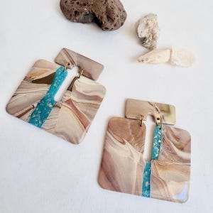 May include: A pair of square earrings featuring a marbled pattern in shades of beige, brown, and cream, with a central vertical strip of bright turquoise blue containing gold flecks. The earrings are connected by small gold-toned metal jump rings.