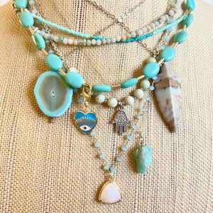 May include: A layered turquoise and white beaded necklace with various charms, including a large agate pendant, a heart-shaped evil eye charm, a hamsa hand charm, and a small triangular pendant.