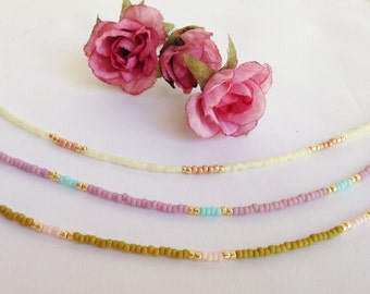May include: Three delicate beaded necklaces with pastel colored beads and gold accents. The necklaces are arranged in a layered fashion on a white background.