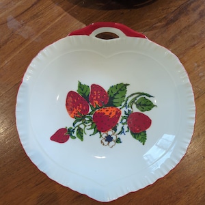 May include: White heart-shaped serving dish with a red handle and rim. The dish features a vibrant strawberry design with green leaves and a small blue flower. The rim has a scalloped edge.