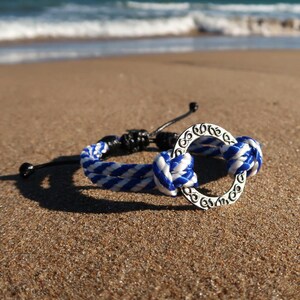 Puede incluir: Una pulsera trenzada azul y blanca con un dije circular plateado, sobre una playa arenosa. La pulsera presenta un diseño náutico, con un patrón trenzado y un dije decorativo. El océano es visible en el fondo.