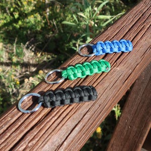 May include: Three paracord keychains in blue, green, and black, each with a silver metal keyring. The keychains are displayed on a weathered brown wooden surface, with a blurred background of greenery.