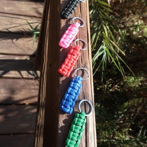 May include: Five colorful paracord keychains in black, pink, red, blue, and green. Each keychain features a woven design and a silver-colored split ring for attaching keys. The keychains are displayed on a wooden surface.
