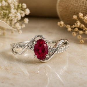 Könnte beinhalten: Ein silberner Ring mit einem zentralen, oval geschliffenen, tiefroten Edelstein, der von vier Krappen gehalten wird. Die Ringschiene hat ein geschwungenes Design mit zwei Armen, die jeweils mit kleinen klaren Steinen besetzt sind.