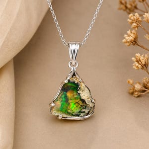 Op de afbeelding: Een zilveren hangerketting met een ruwe, driehoekige opaal edelsteen met levendige groene en oranje kleurflitsen. De steen wordt vastgehouden in een gepolijste zilveren zetting met pootjes en hangt aan een fijne zilveren schakelketting.