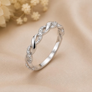 Puede incluir: Un anillo de tono plateado con un diseño de banda trenzada. Una mitad de la banda trenzada es pulida y lisa, mientras que la otra mitad está incrustada con pequeñas piedras redondas y transparentes que captan la luz, creando un efecto brillante.