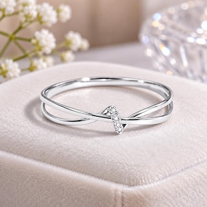 May include: A silver-coloured ring featuring a delicate crossover band design. The centre of the ring showcases a small, vertical bar element encrusted with four tiny, clear, sparkling stones that catch the light, creating a refined and polished appearance.
