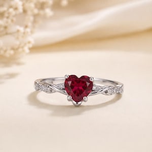 Puede incluir: Un anillo de tono plateado con una piedra preciosa central de color rojo intenso en forma de corazón, sujeta por garras. La banda presenta un diseño trenzado y está adornada con pequeñas piedras claras y brillantes incrustadas en los laterales.