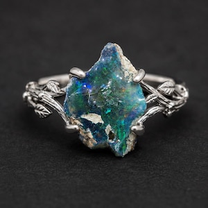May include: A silver ring featuring a raw, uncut opal gemstone with vibrant blue and green flashes. The band is designed with a detailed, textured vine and leaf motif that extends from the central setting, securing the stone with four prongs.
