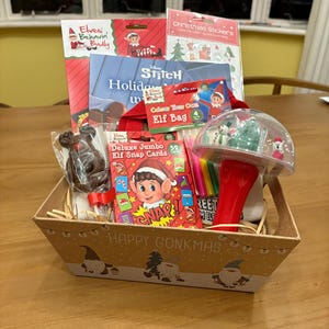 Pode incluir: Uma cesta de presente cheia de itens com tema de Natal. Inclui um livro, adesivos, um livro Disney Stitch, uma sacola de Elfo, cartões Elf Snap, uma rena de doces e um brinquedo tipo globo de neve. A cesta é decorada com gnomos e o texto "HAPPY GONKMAS".